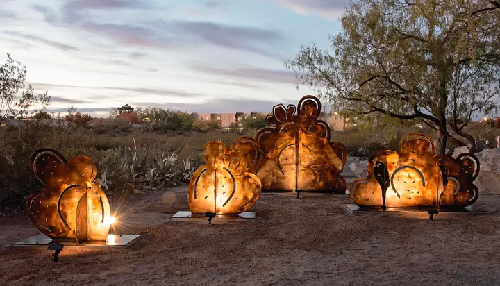 Metal sculpture panels shaped like desert plants glow from internal lighting at dusk in a landscaped Las Cruces park, with trees, cacti and buildings visible in the background.