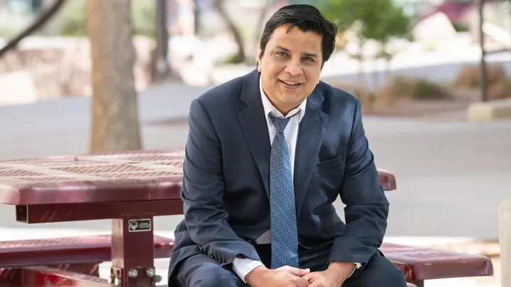 Jagdish Khubchandani, in a navy suit and tie, sits at an outdoor picnic table and smiles toward the camera on the New Mexico State University campus.