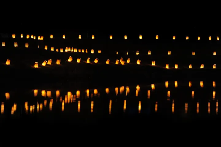 Rows of glowing luminarias line a dark pathway at night, their warm light reflecting off the water below.