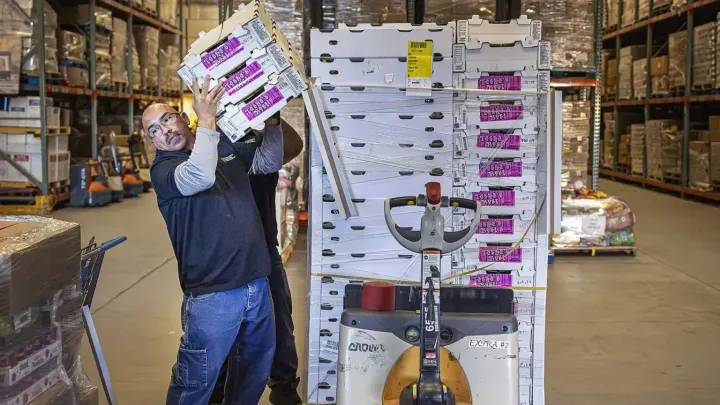 A worker lifts stacked produce boxes onto his shoulder inside a warehouse, standing next to a pallet jack loaded with shrink-wrapped cartons, with shelves of food visible in the background.