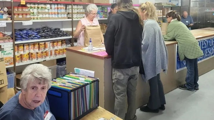 Volunteers at a food pantry prepare bags of groceries for people waiting at the counter, with shelves of canned goods and staples visible behind them.