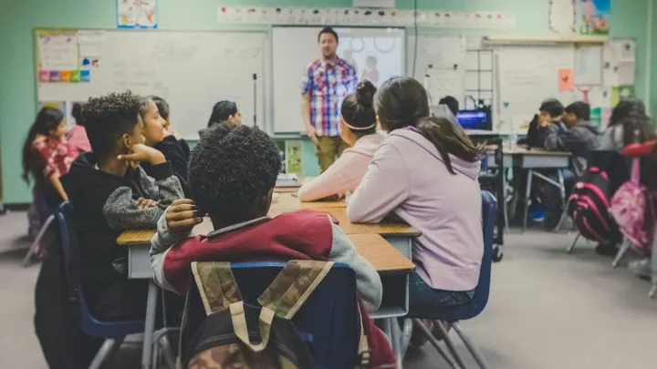 A classroom full of students sits at desks facing a teacher at the front of the room during a lesson.