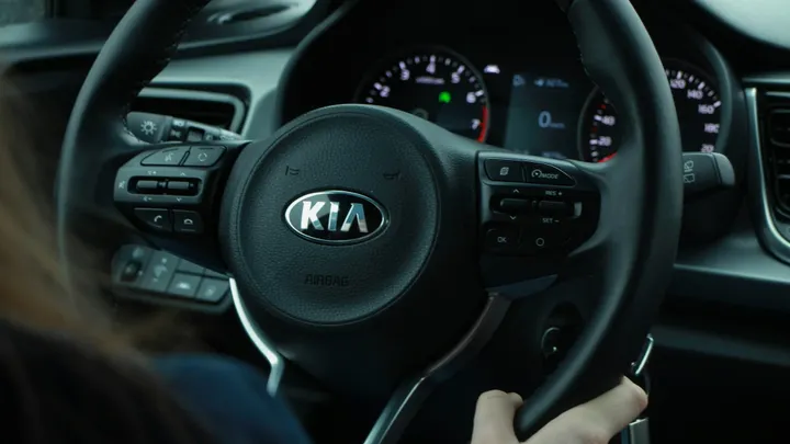 Driver’s view of a Kia vehicle interior, with hands on the steering wheel and the Kia logo visible at the center of the wheel.