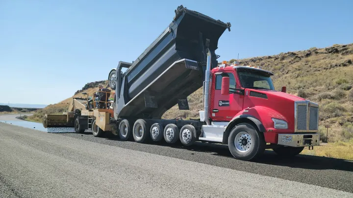 A dump truck unloads asphalt as road construction equipment repaves a highway in a rural area.