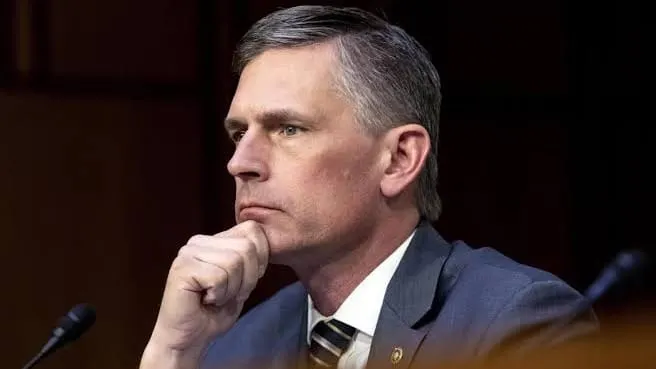 U.S. Sen. Martin Heinrich sits at a hearing table with his hand on his chin, wearing a gray suit and striped tie, with microphones and a blurred background behind him.