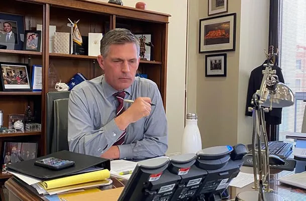 U.S. Sen. Martin Heinrich sits at his desk in his office, holding a pen near his mouth while looking at a row of desk phones, with framed photos, books and a suit jacket visible behind him.