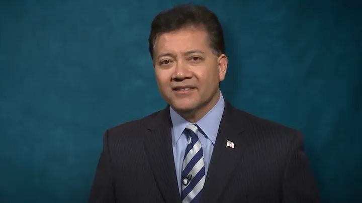 Ken Miyagishima wearing a dark suit and striped tie speaks on camera against a solid blue studio backdrop.