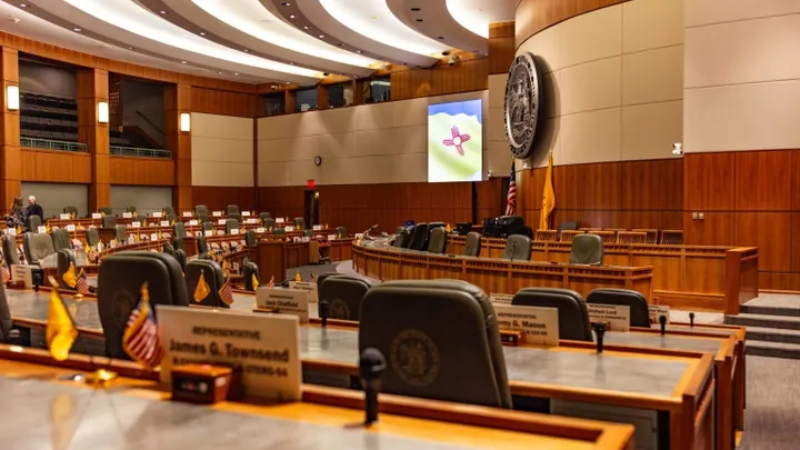 Empty New Mexico House chamber in Santa Fe, with curved rows of desks, nameplates, small U.S. and state flags, the state seal on the wall and the New Mexico flag displayed on a screen.