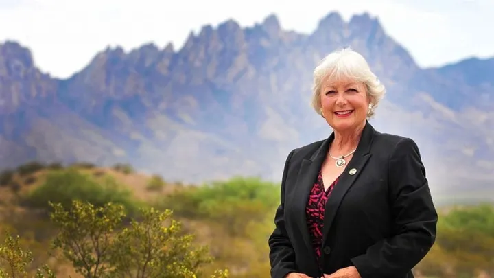 State Rep. Joanne Ferrary stands outdoors in front of the Organ Mountains, wearing a black blazer and patterned top, with desert vegetation visible in the foreground.