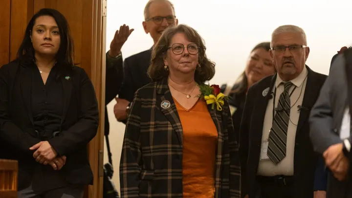 State Sen. Linda Trujillo stands indoors with other officials in a legislative building hallway, wearing an orange top and plaid jacket, as people gather behind her.