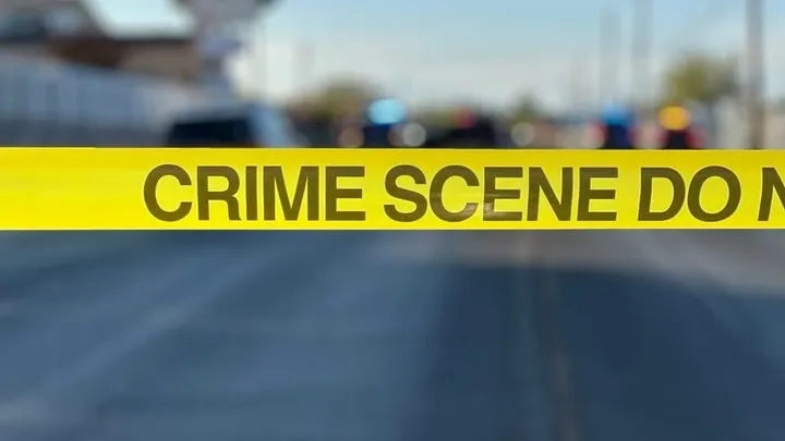 Yellow crime scene tape reading “Crime Scene Do Not Cross” stretches across a street with police vehicles blurred in the background.