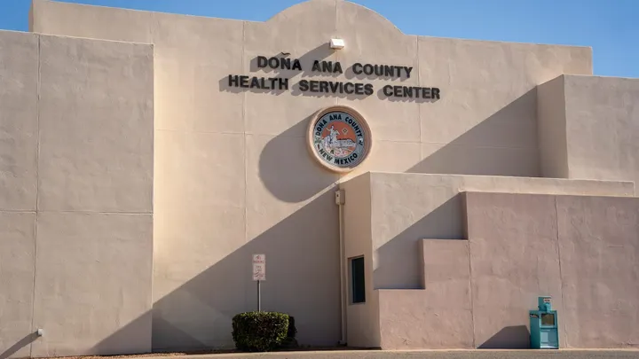 Exterior of the Doña Ana County Health Services Center building in Las Cruces, with the county seal mounted on the stucco facade beneath a clear blue sky.