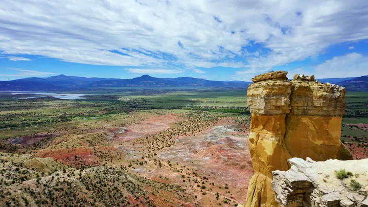 Colorful desert landscape with layered rock formations and mesas overlooking a wide New Mexico valley under a partly cloudy sky.