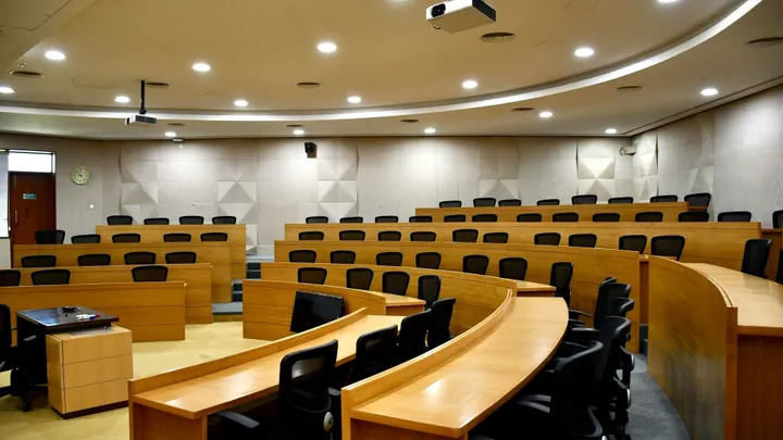 Empty courtroom with tiered seating, wood-paneled desks and a judge’s bench at the front.
