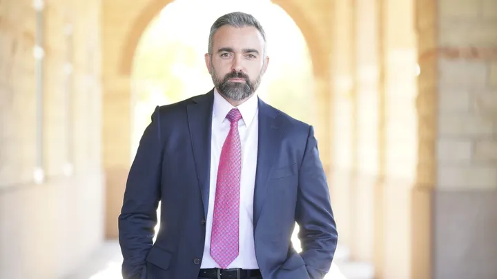 New Mexico Attorney General Raúl Torrez in a suit and tie standing in an arched corridor.