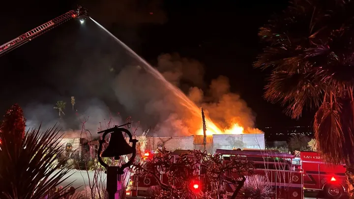 Firefighters spray water from a ladder truck onto a house fire at night, with flames and heavy smoke rising from the structure in the Las Alturas area.