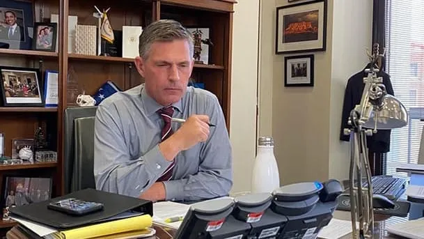 U.S. Sen. Martin Heinrich sits at his desk in an office, holding a pen during a call, with framed photos and bookshelves visible behind him.