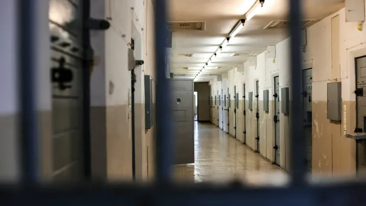 Interior of a prison cellblock hallway lined with closed metal doors, viewed through bars