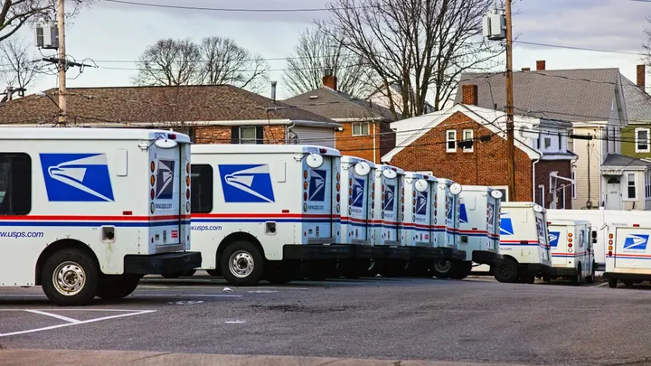 U.S. Postal Service delivery trucks are lined up in a parking lot near a residential neighborhood