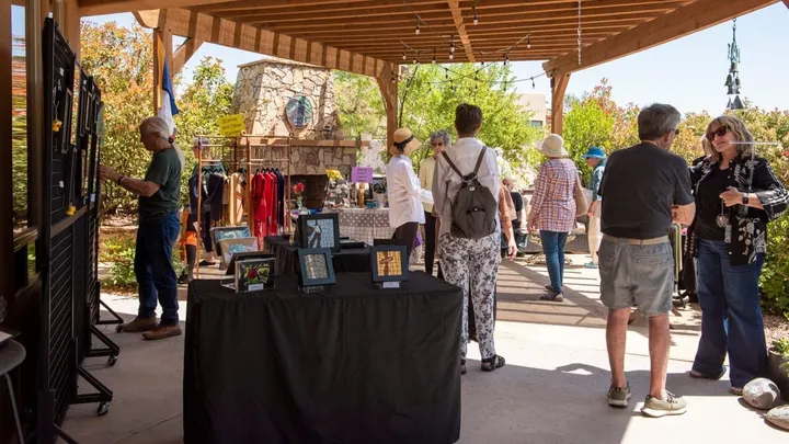 People walking through an outdoor garden art show, viewing artwork displayed on tables and stands under a covered patio.