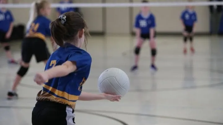 Youth volleyball player serving the ball during an indoor game while teammates prepare on the court