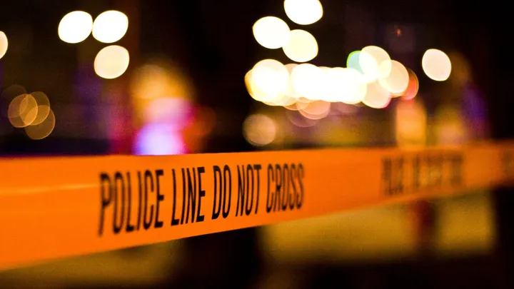 Police tape reading “Police Line Do Not Cross” stretched across a street at night with blurred emergency lights in the background