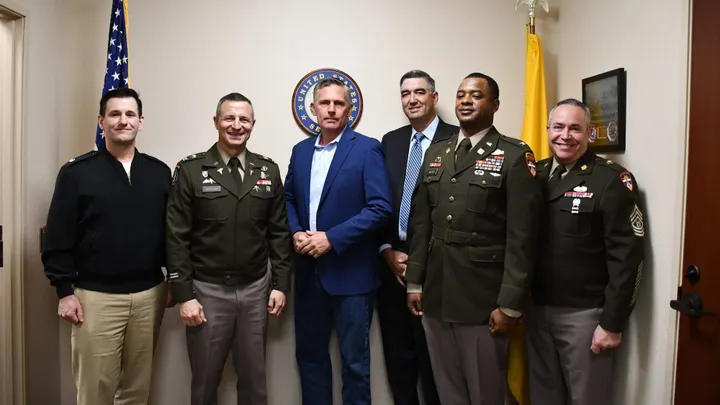 U.S. Sen. Martin Heinrich stands with White Sands Missile Range leadership and military personnel in a group photo inside an office, with U.S. and New Mexico flags in the background.