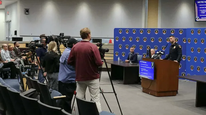 Police officials speak at a podium during a news conference as media members record and take photos in a conference room.