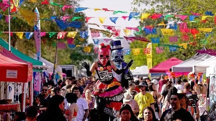 Festival crowd in downtown Las Cruces with colorful banners and costumed performers walking above attendees on stilts.