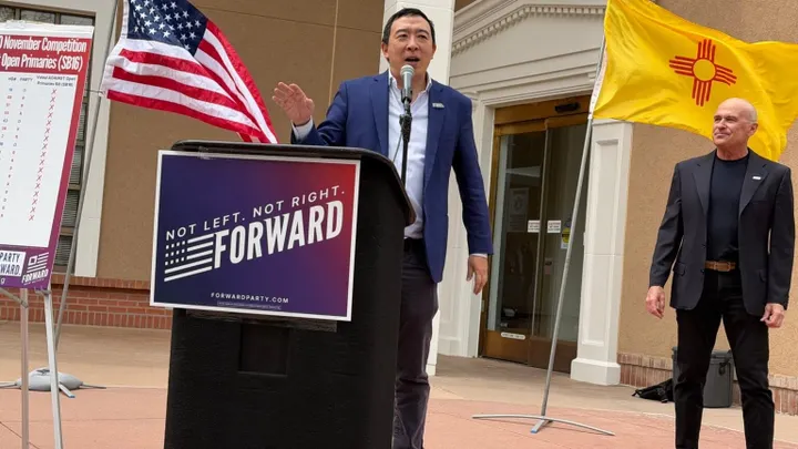 Andrew Yang speaks at a podium during a Forward Party event in New Mexico, with U.S. and state flags displayed behind him.