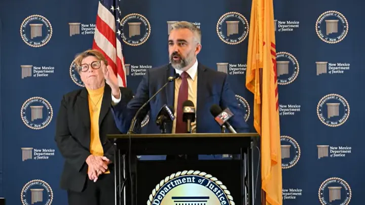 New Mexico Attorney General Raúl Torrez speaks at a podium during a press conference, with a woman standing behind him against a New Mexico Department of Justice backdrop.