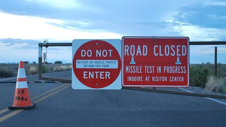 Roadblock on a highway with signs reading “Do Not Enter” and “Road Closed — Missile Test in Progress,” along with a traffic cone blocking access.