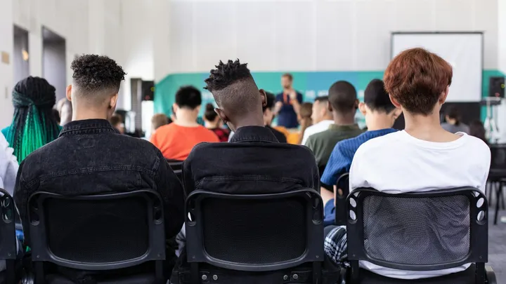 Students seated in a classroom or assembly listening to a speaker at the front of the room