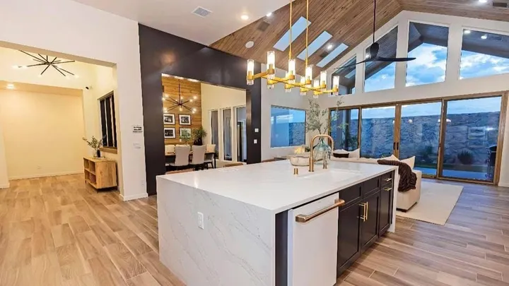 A modern open-concept kitchen and living area features a large white island, wood-paneled ceiling accents, pendant lighting and floor-to-ceiling windows overlooking a backyard patio.
