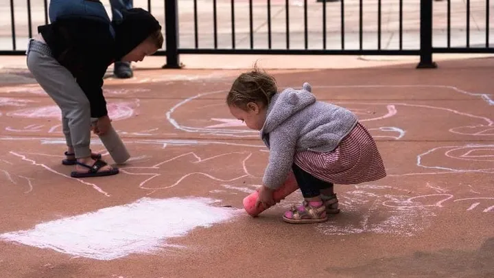 Two young children crouch on a sidewalk while drawing with colorful chalk near a metal fence.