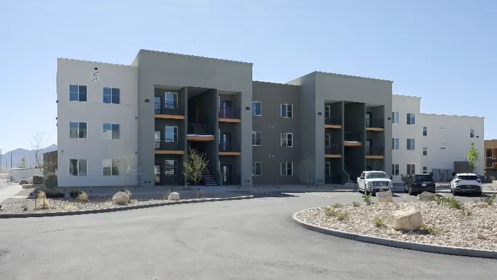 A newly constructed multi-story apartment building stands beside a curved driveway under a clear sky in Las Cruces.
