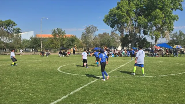 Children play soccer on a grassy field during a youth outdoor league game in Las Cruces while spectators watch from the sidelines.