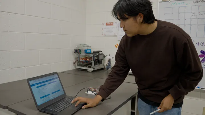 Student works on a laptop connected to a small electronic sensor device during a demonstration in a school STEM lab.