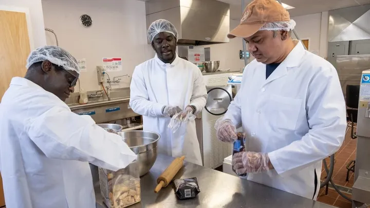 Three New Mexico State University graduate students wearing lab coats, hairnets and gloves prepare ice cream ingredients on a stainless steel worktable in the campus food science dairy lab.