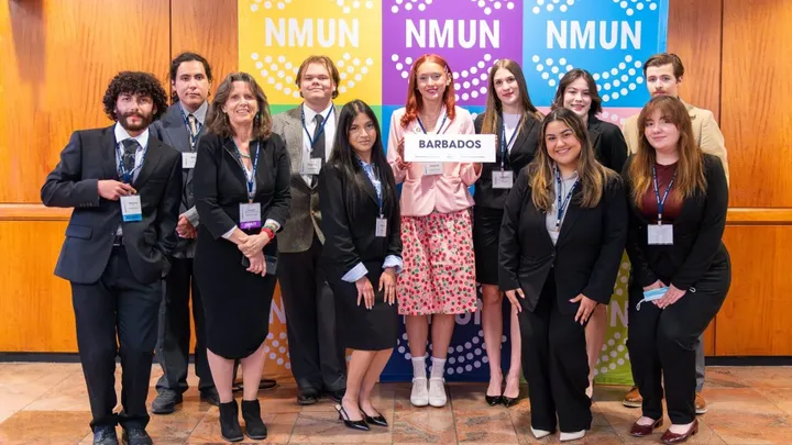 Group of New Mexico State University Model United Nations students and faculty posing at a conference, with one student holding a sign labeled “Barbados” in front of NMUN banners.