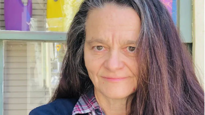 Connie Voisine, a poetry professor at New Mexico State University, stands outdoors in front of a window while wearing a plaid shirt and dark cardigan.