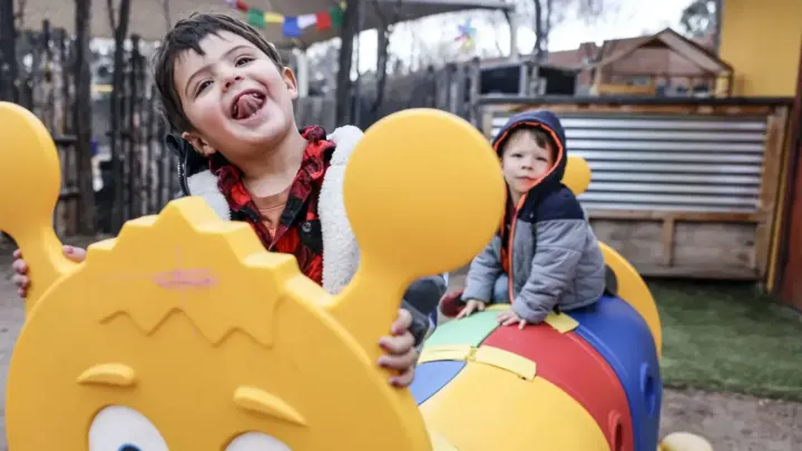 Two young children play on colorful playground equipment at a childcare center, with one child smiling toward the camera.