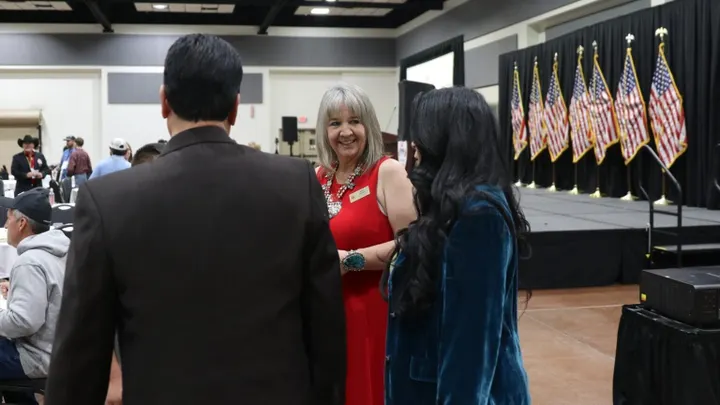 Amy Barela, chair of the New Mexico Republican Party, speaks with attendees at an indoor event with American flags in the background.