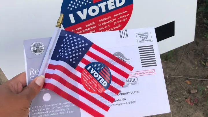 An absentee ballot envelope, small American flag and “I Voted” stickers are displayed in front of a ballot drop box.