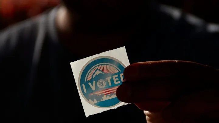A person holds an “I Voted” sticker in front of a dark background.