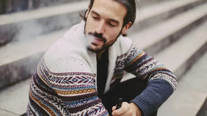 A man sits on outdoor steps while exhaling vapor from an e-cigarette.