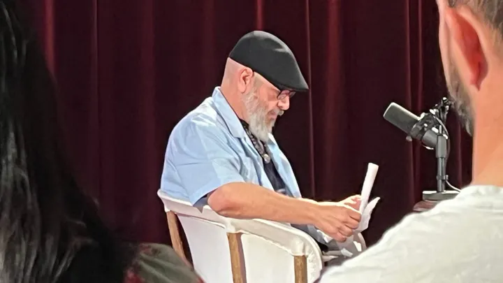 Manuel González, wearing a black cap and light blue shirt, looks down at papers in his hands while seated beside a microphone before a live “Pass the Sopapillas” taping at KRWG Studios.