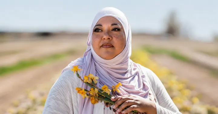 Daisy Maldonado wears a pale pink hijab and holds yellow flowers in an outdoor portrait with a softly blurred landscape in the background.