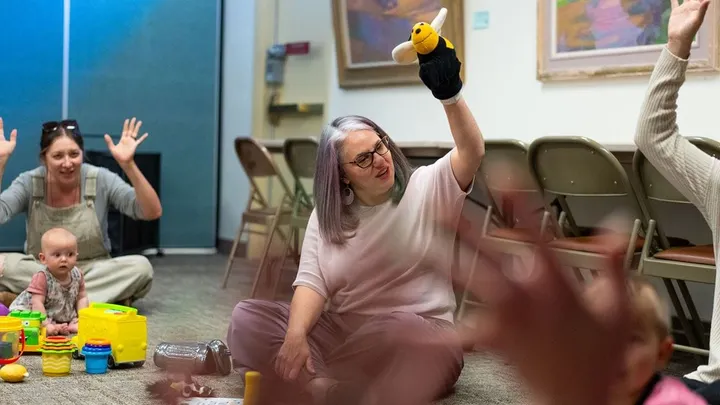 Nancy Stephens holds up a bee puppet while sitting on the floor with children and adults during an Imagination Library event.