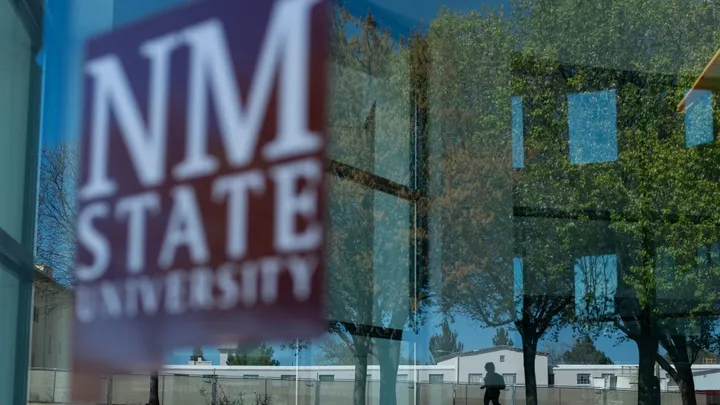 Blurred New Mexico State University sign reflected in a window with trees and campus buildings visible outside.